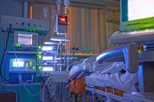 Hospital intensive care unit (ICU) room showing a patient in bed connected to advanced life support monitors, infusion pumps, and multiple medical devices glowing in dim lighting.