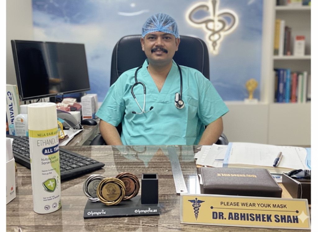 Doctor in green scrubs and surgical cap, with a stethoscope around the neck, sits at a desk with medals, sanitizer, paperwork, and a nameplate reading "Dr. Abhishek Shah."