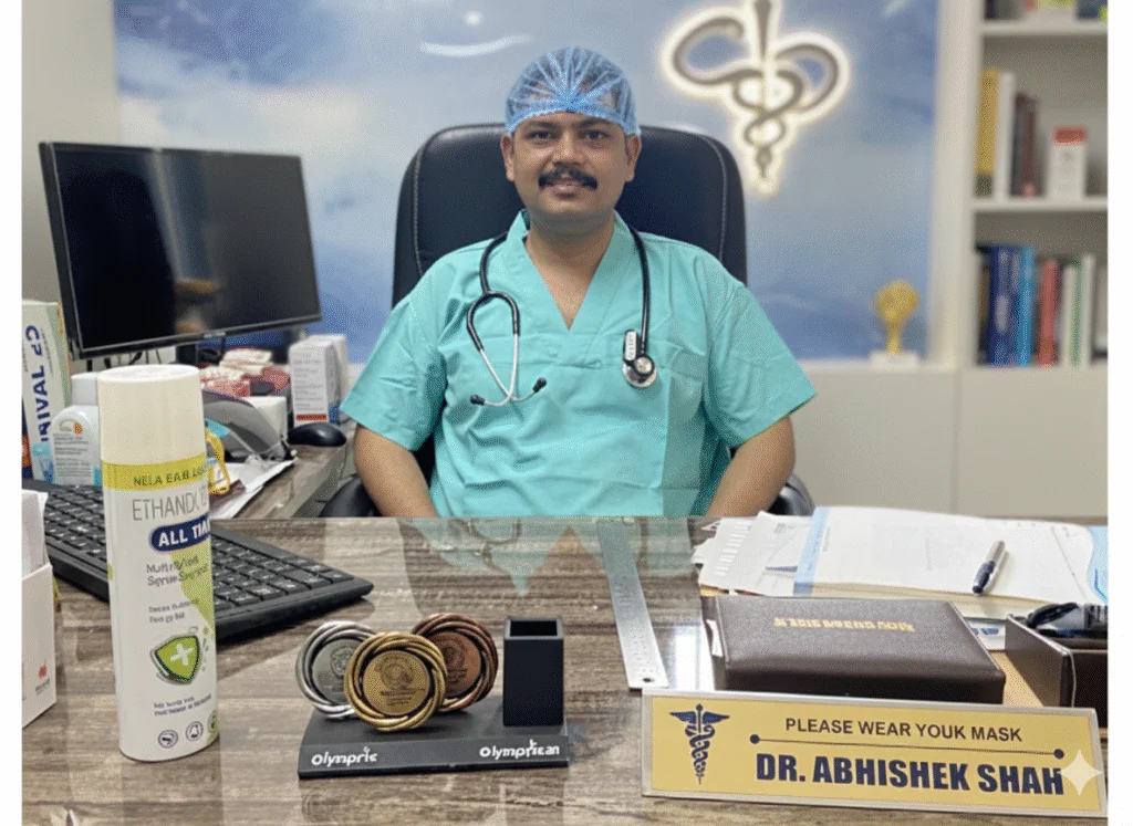 Doctor in green scrubs and surgical cap, with a stethoscope around the neck, sits at a desk with medals, sanitizer, paperwork, and a nameplate reading "Dr. Abhishek Shah."