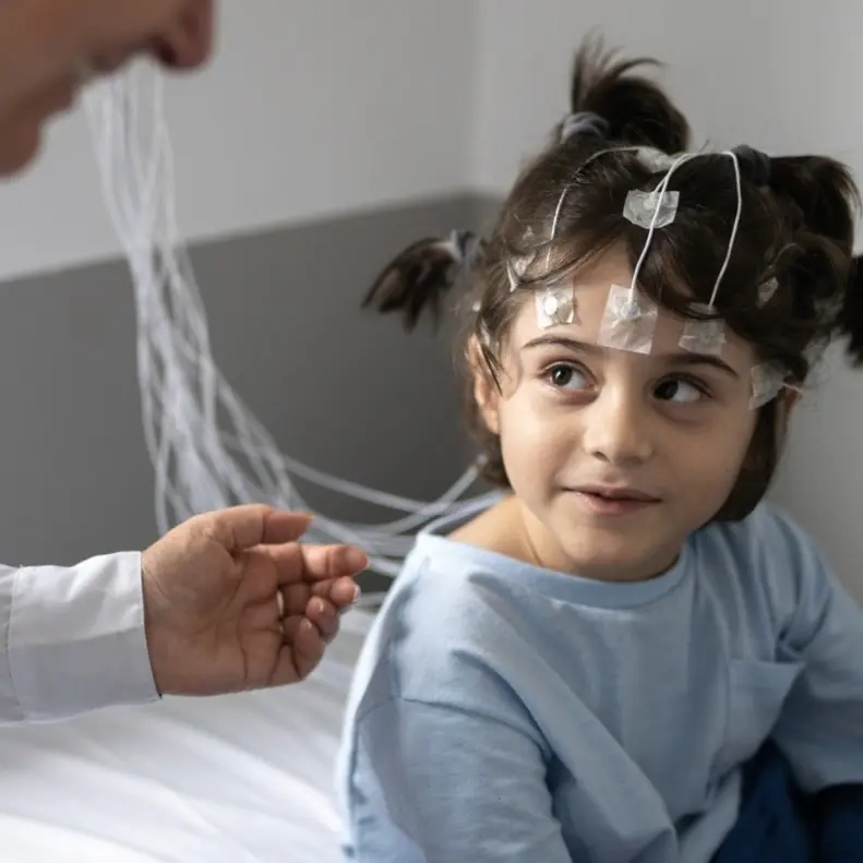 Young girl with EEG electrodes and wires attached to her head, sitting in a hospital setting with a doctor nearby, preparing for a neurological test.