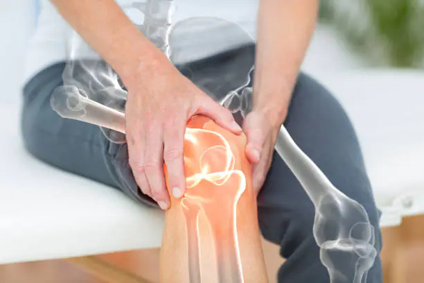 Person seated on an examination table holding their sore knee, with a transparent overlay of bones and joint, highlighting pain and anatomy.