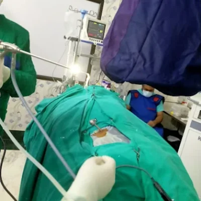 Surgeons performing a minimally invasive procedure on a patient draped in green in an operating room, using endoscopic instruments, with a medical team member monitoring equipment in the background.