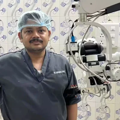 Surgeon in gray scrubs and a blue hair cap stands beside a large operating room microscope or robotic surgical device, with patterned tiles on the wall in the background.